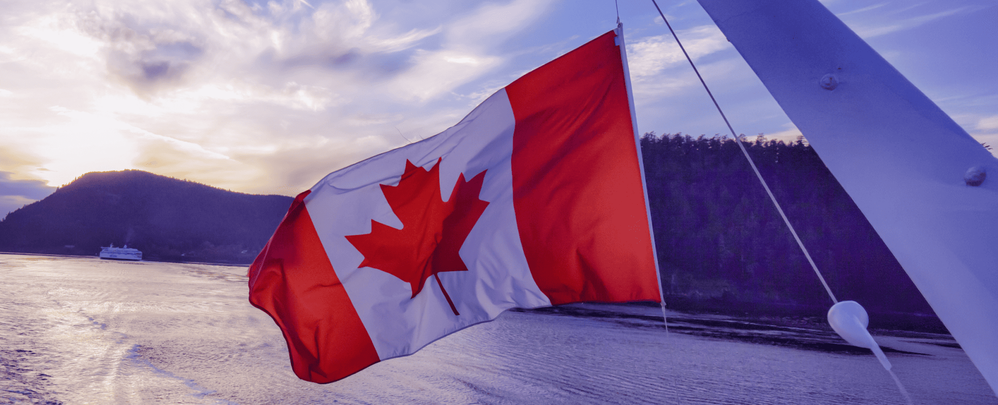 A Canadian flag waving with a body of water and forested hills in the background at sunset.