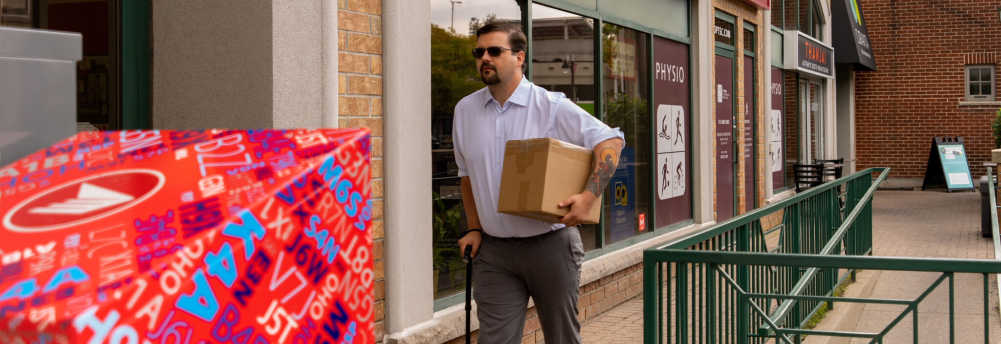A man carrying a package walks with an assistive cane on a sidewalk ramp along storefronts. 