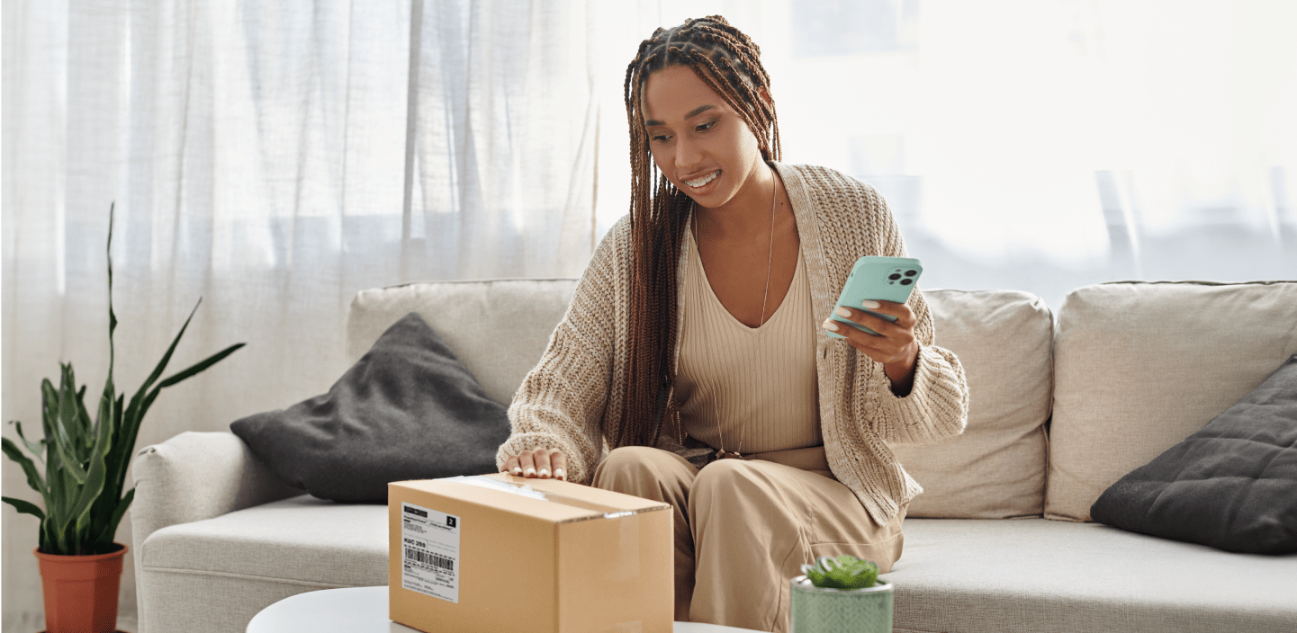 A smiling woman looks at her smart phone as she holds a package
