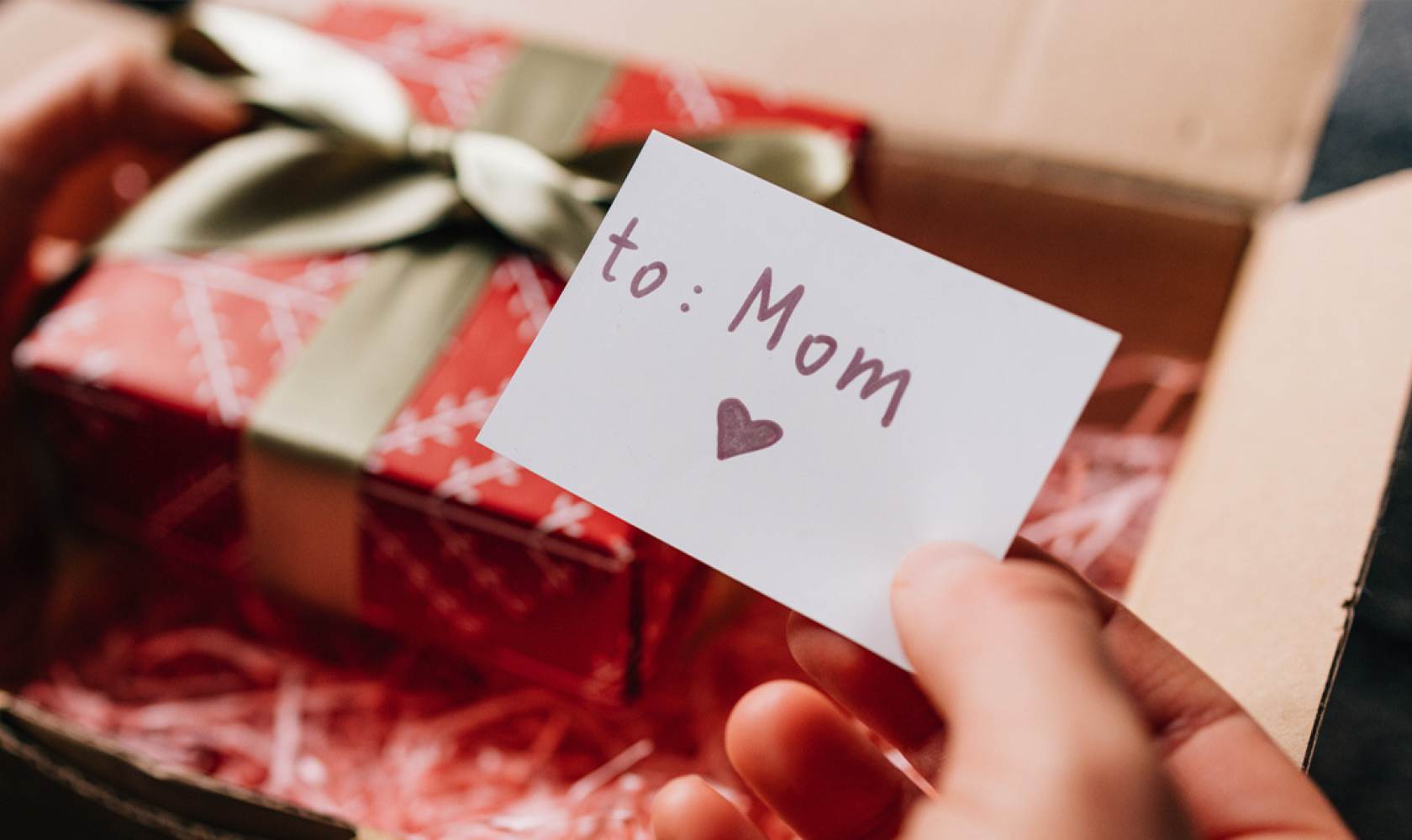 Hands open a shipping box which contains a wrapped gift box and a card that reads 'to Mom.’