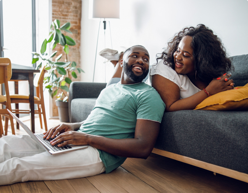 A man types on a laptop and leans against a couch a woman is sitting on. They smile at each other. A man types on a laptop and leans against a couch a woman is sitting on. They smile at each other.