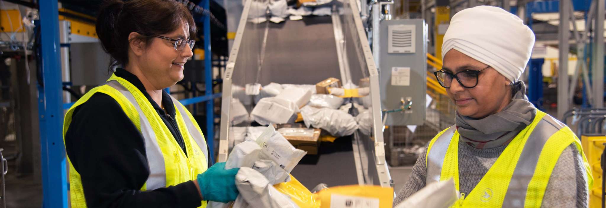 Two female Canada Post employees wearing bright yellow safety vests sort through packages and envelopes.