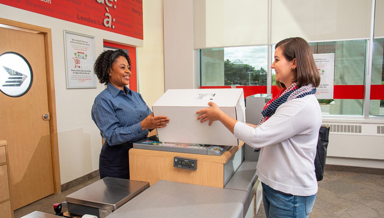 A smiling Canada Post employee hands a package to a smiling customer at a post office.
