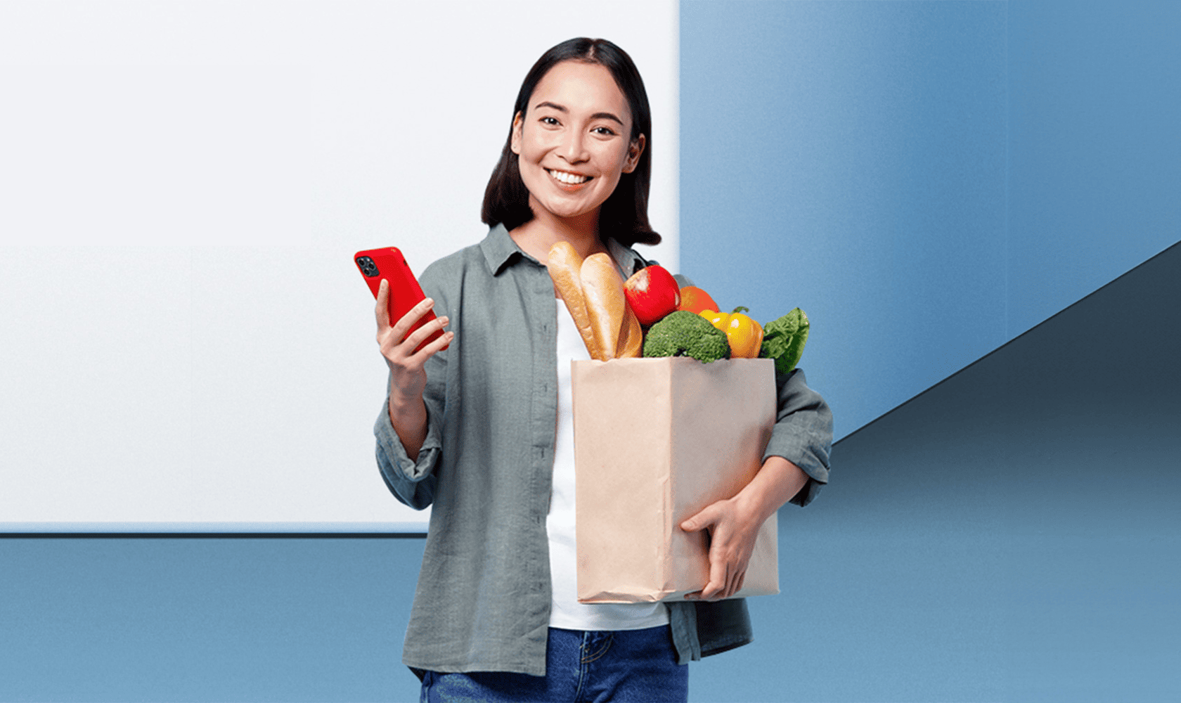 A woman smiles and holds a cell phone in one hand and a bag of groceries in the other.