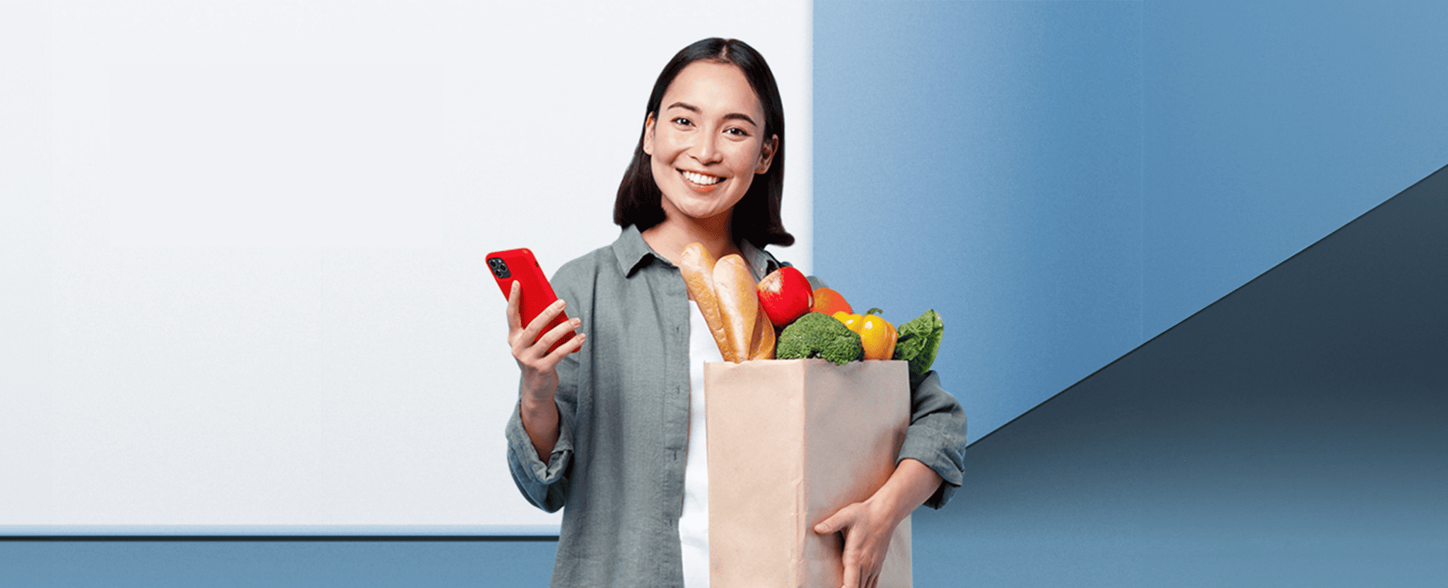A woman smiles and holds a cell phone in one hand and a bag of groceries in the other.
