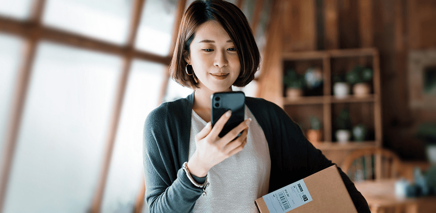 A woman looks at her mobile phone and smiles, holding a package with a return label under her arm.