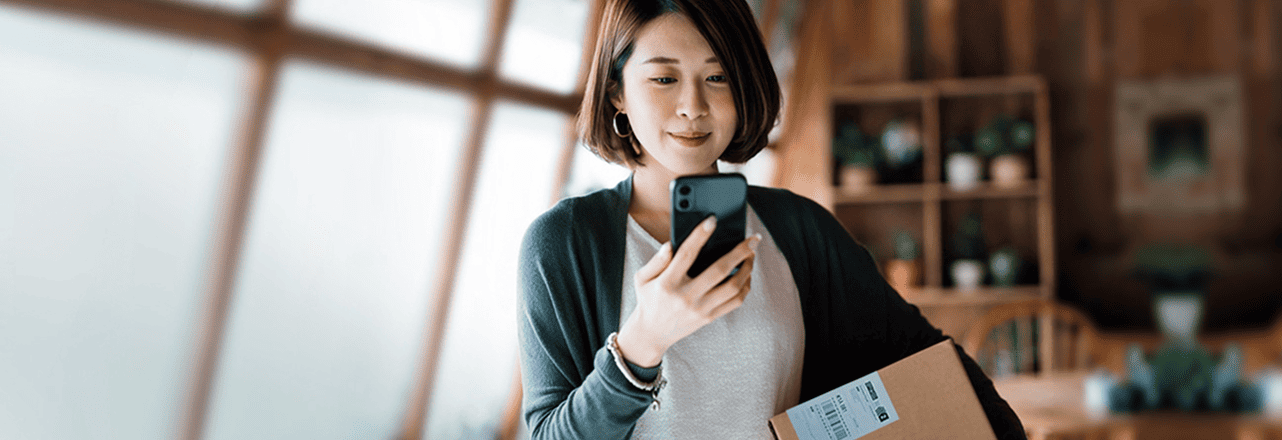 A woman looks at her mobile phone and smiles, holding a package with a return label under her arm.