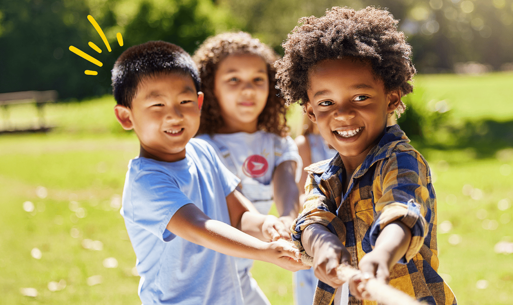Children playing tug-of-war outdoors, pulling a rope together on a sunny day with grass and trees in the background.