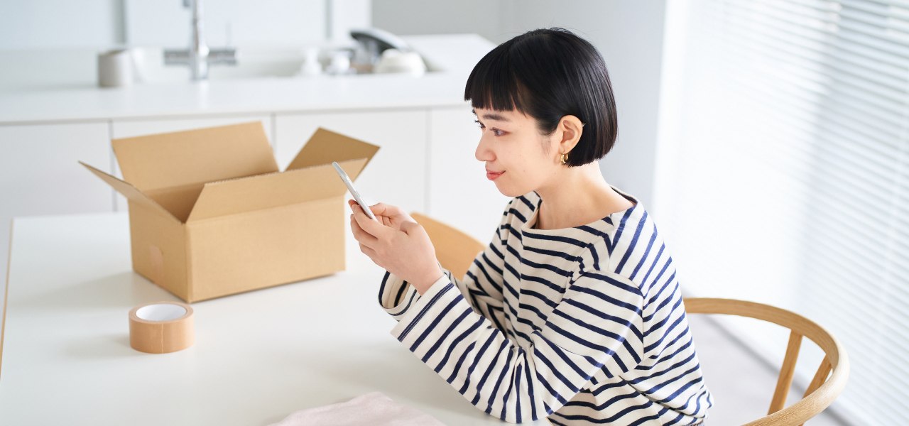 A woman types on her mobile phone. She sits at a table that has an open shipping box and a roll of tape on top.