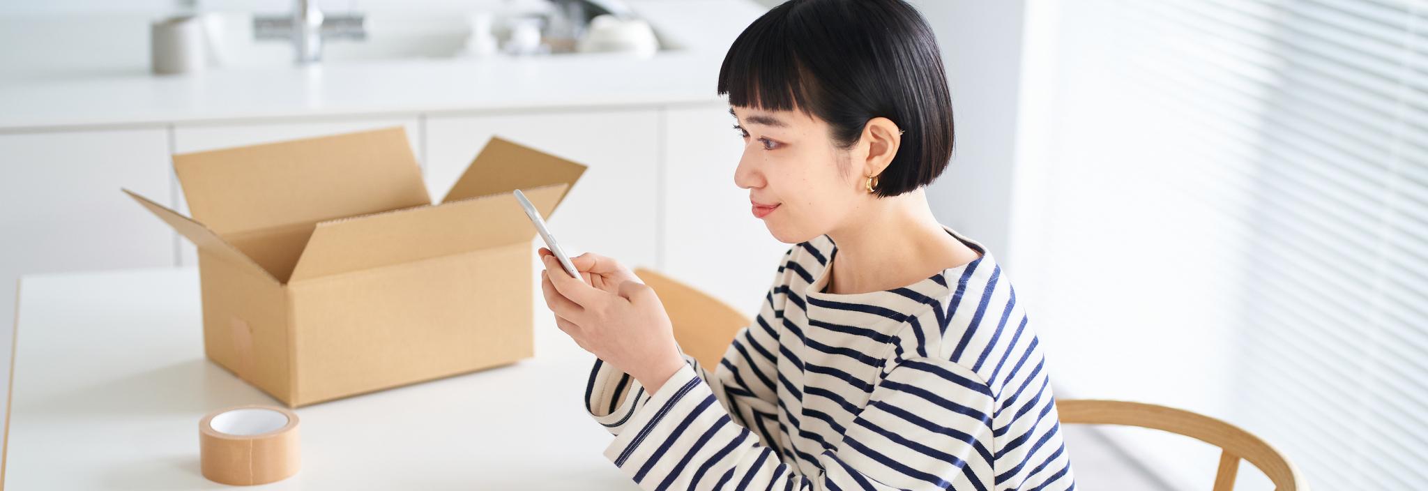 A woman types on her mobile phone. She sits at a table that has an open shipping box and a roll of tape on top.