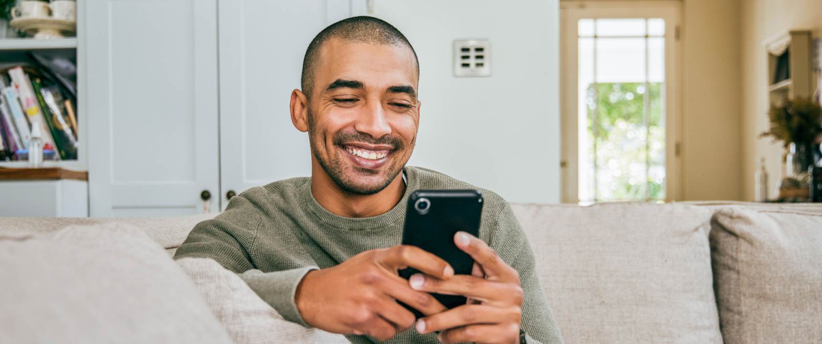 A young man smiles and stares a cellphone in his hands. He sits on a couch in his living room.