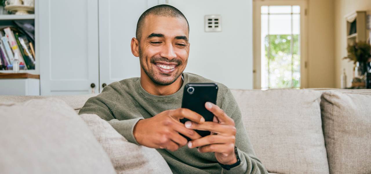 A young man smiles and stares a cellphone in his hands. He sits on a couch in his living room.