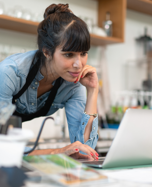 A woman in an apron leans forward and types on a laptop sitting on the service counter in a cafe.