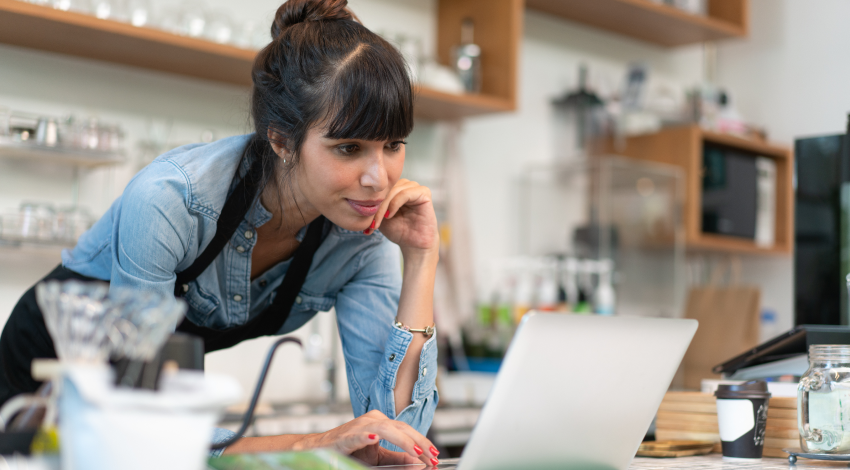 A woman in an apron leans forward and types on a laptop sitting on the service counter in a cafe.