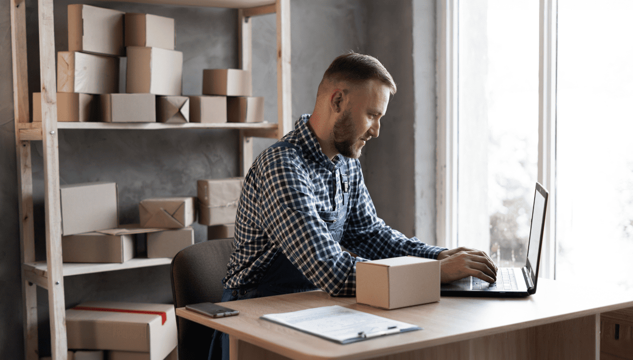 A man sits at a desk and types on a laptop in a room full of boxes.