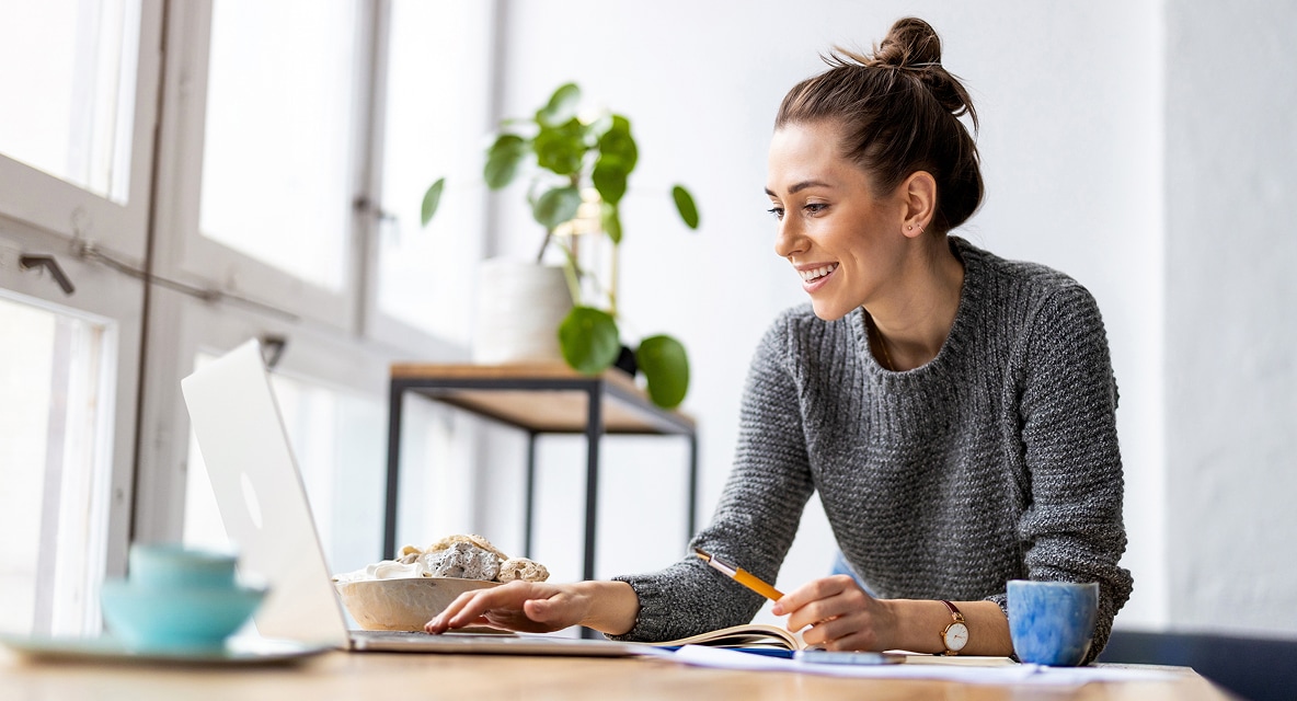 A smiling woman sits at a table in a bright, sunlit room, working on a laptop.