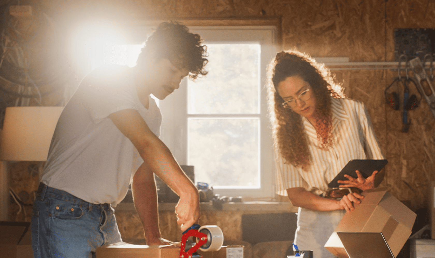 People packing a cardboard box in a sunlit room, with one using tape and another holding a tablet.