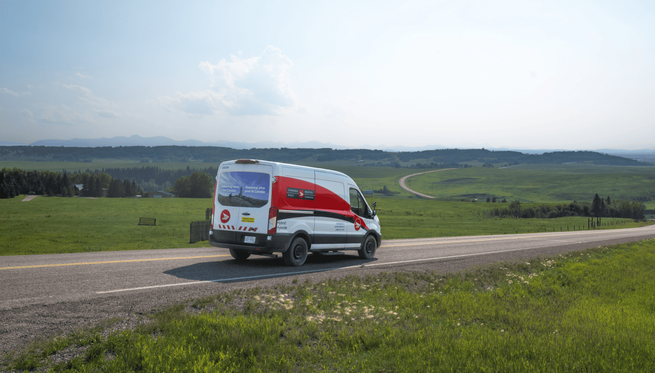 Canada Post delivery van driving along a rural highway through rolling green hills under a hazy sky.