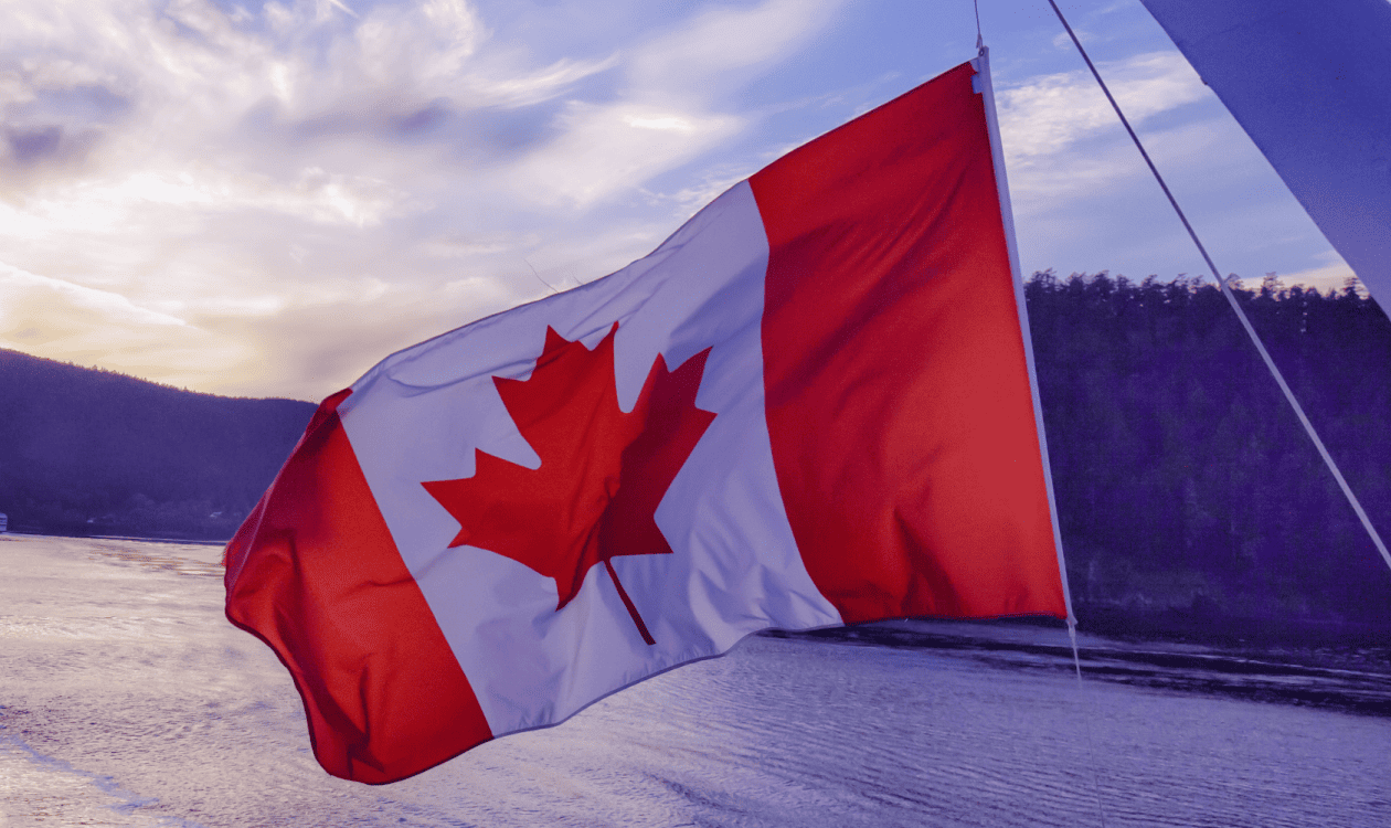 A Canadian flag waving with a body of water and forested hills in the background at sunset.