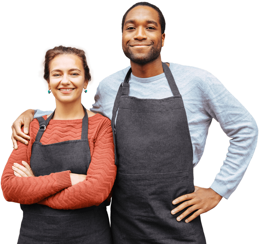 A smiling man wears an apron. His arm is around a woman wearing an apron.