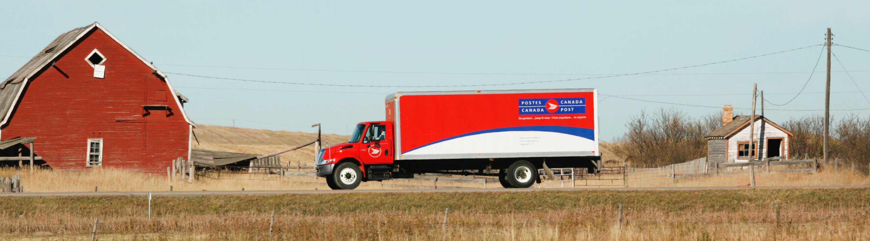 A Canada Post truck drives towards a barn on a rural road.