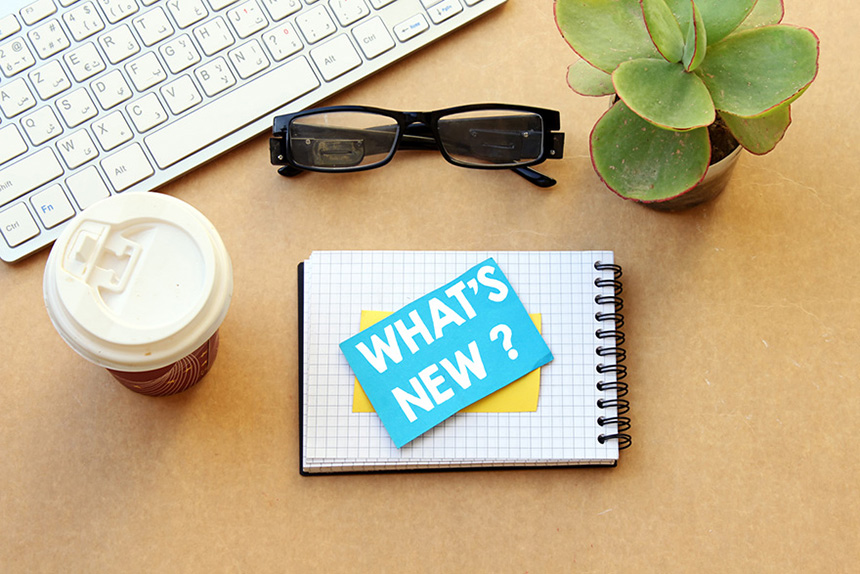 A birds eye view shows different objects on a table : a keyboard, a little plant, a pair of glasses, a coffee and a notebook with a yellow Post-it note on it reading “WHAT’S NEW?”