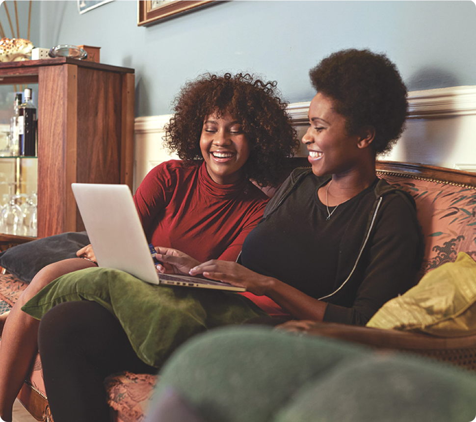 Two women sit together on a couch and look at a laptop screen.