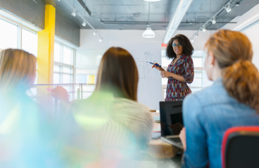 A standing woman presents to three seated women in an office boardroom.