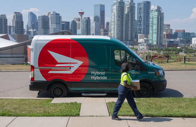 A Canada Post driver walks on a sidewalk and carries a package for delivery. A hybrid Canada Post van is parked behind her on the street. In the background is the Calgary skyline.