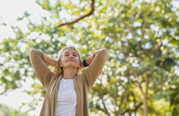 A mature woman runs her hands through her hair as she walks through a heavily treed area.