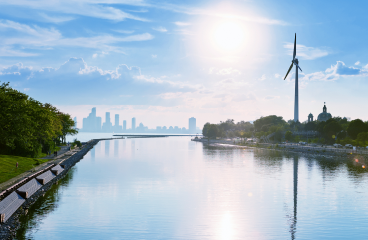 Une grande éolienne surplombe une rivière et du côté opposé, des gratte-ciel s’élèvent dans la brume au loin