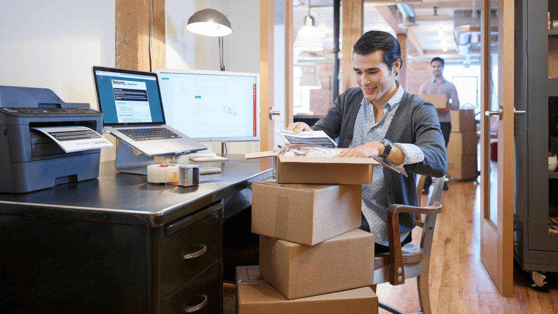 A smiling man sits at a desk in an office. He packs a shipping box that is stacked on top of other shipping boxes.