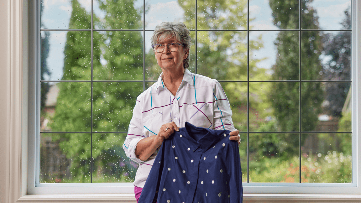 Catharine Janssen, propriétaire de BRACAShirts, se tient devant une grande fenêtre. Elle tient une tunique à motifs qu’elle a conçue et fabriquée.