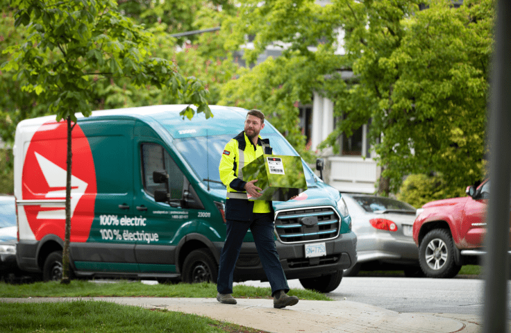 A Canada Post driver stands in front of an electric Canada Post van. He carries a cardboard package for delivery.