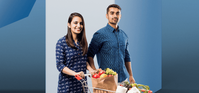 A smiling couple push a grocery cart filled with items.
