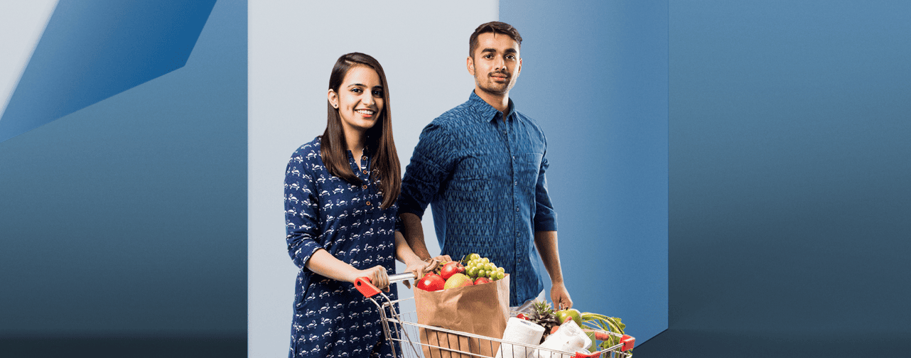 A smiling couple push a grocery cart filled with items.