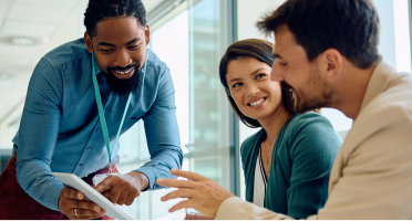 Trois personnes collaborent dans un bureau moderne, l’une se tient debout avec une tablette tandis que les autres sont assises.