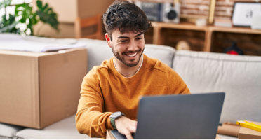 Un homme assis sur un canapé sourit devant un ordinateur portable. Il est entourée de boîtes, suggérant un déménagement récent ou une activité de commerce en ligne à la maison.