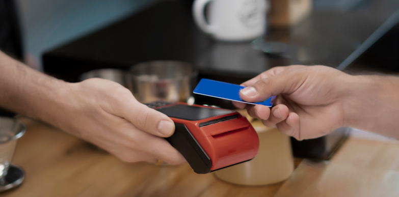 Une main tient un terminal de paiement rouge tandis que la main d'une autre personne se sert d’une carte bleue lors d’une transaction sans contact au comptoir d’un café.