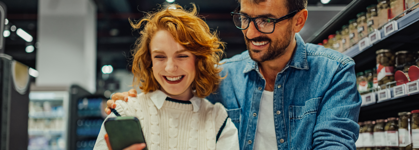 Une femme et un homme derrière un panier d'épicerie consultent un téléphone intelligent, avec des étagères en arrière-plan.