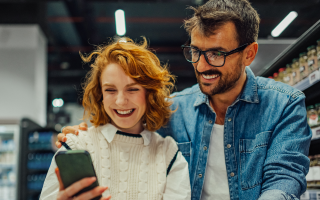 Une femme et un homme derrière un panier d'épicerie consultent un téléphone intelligent, avec des étagères en arrière-plan.