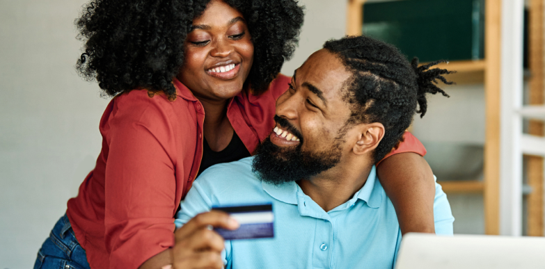 Un homme tient une carte près d’un ordinateur portable tandis qu’une femme se penche sur son épaule. Le couple semble conclure un achat en ligne ou une transaction financière commune.