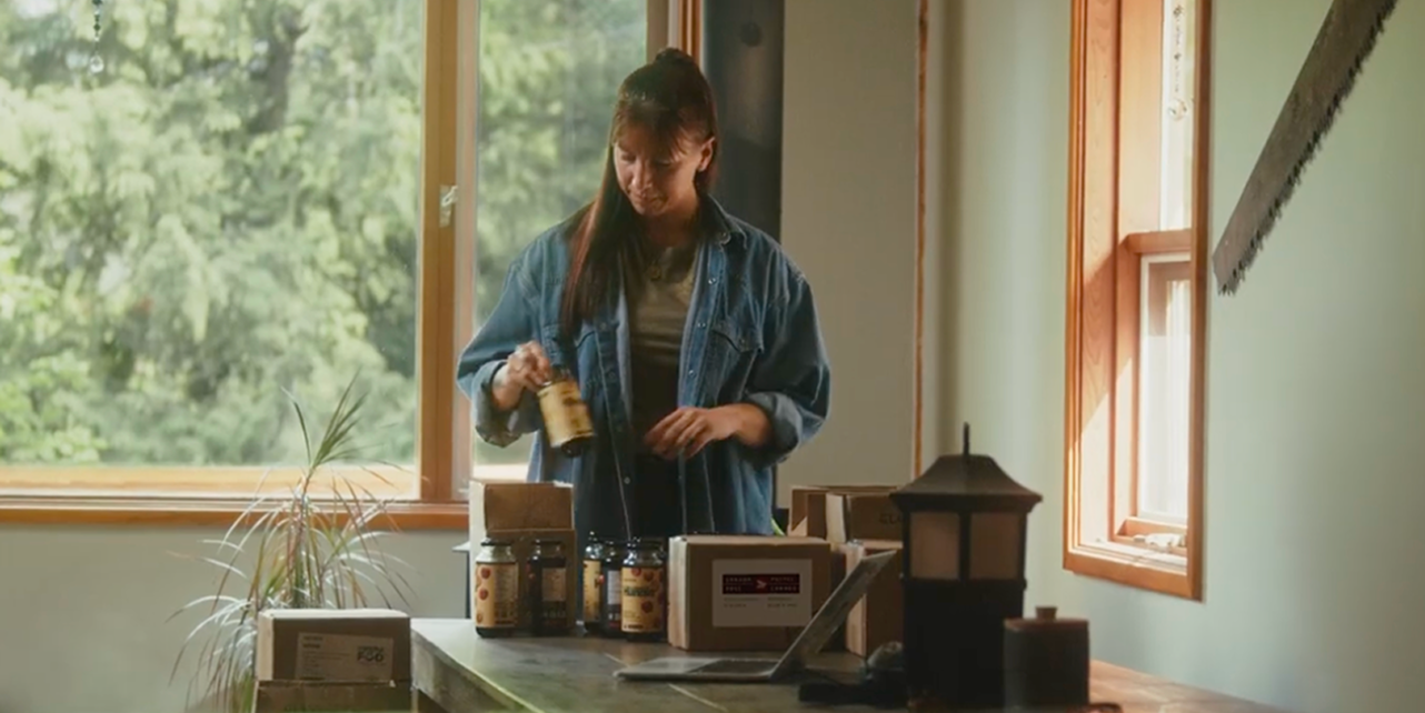 Venessa Stonehouse, cofondatrice et chef de la direction de Mindful Food, place des pots &eacute;tiquet&eacute;s dans des bo&icirc;tes d&rsquo;exp&eacute;dition sur une table, &agrave; c&ocirc;t&eacute; d&rsquo;un ordinateur portable, dans un espace de travail baign&eacute; de lumi&egrave;re naturelle.