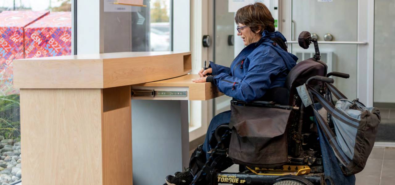 A woman in a wheelchair fills out a form at an accessible desk in a post office. 