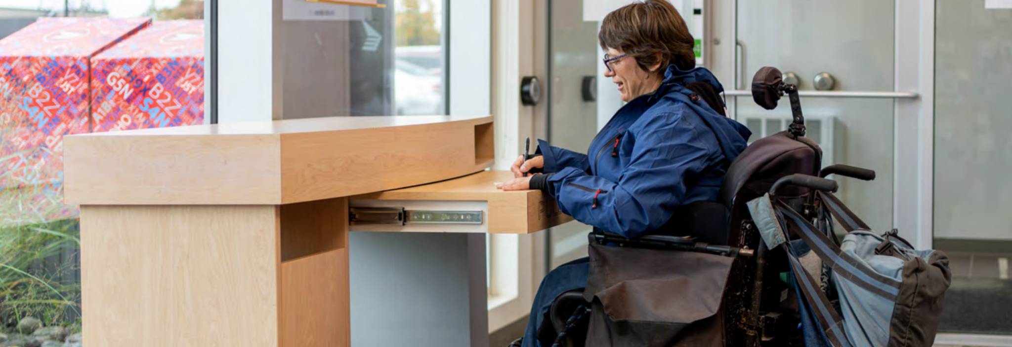 A woman in a wheelchair fills out a form at an accessible desk in a post office. 