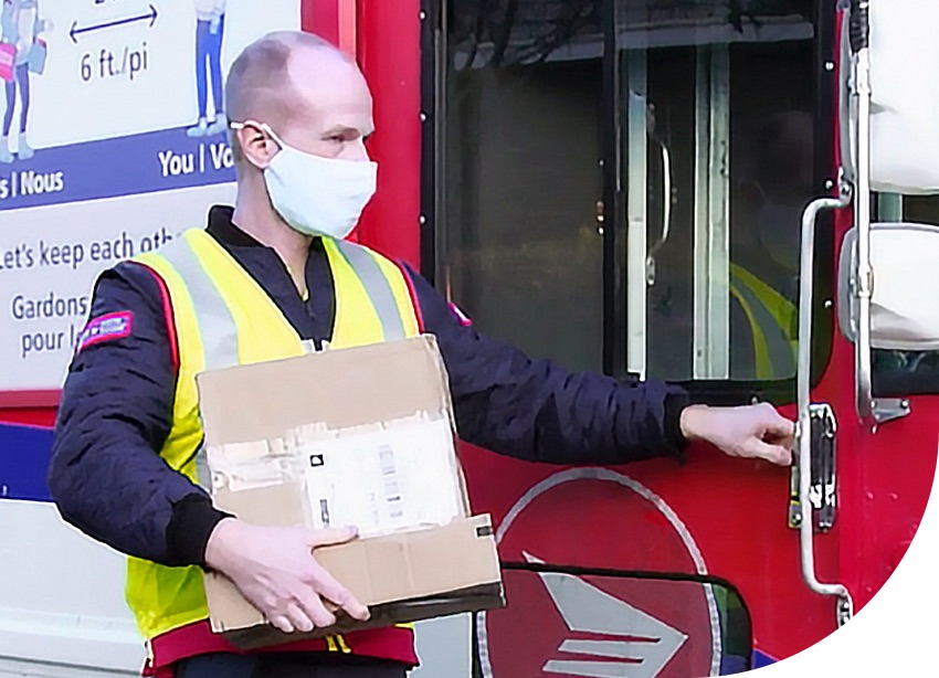 A Canada Post mail carrier wears a mask and holds a parcel beside his delivery vehicle.