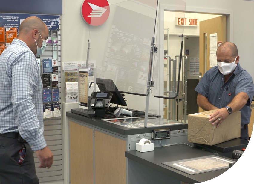 A Canada Post employee wears a mask and processes a parcel behind a counter with a protective barrier in a post office.
