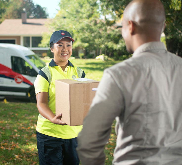 A Canada Post delivery person hands a package to a customer on a lawn.