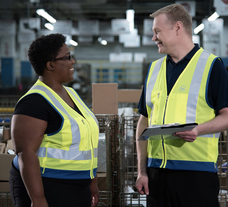 Two Canada Post workers smile at each other in a sorting facility.