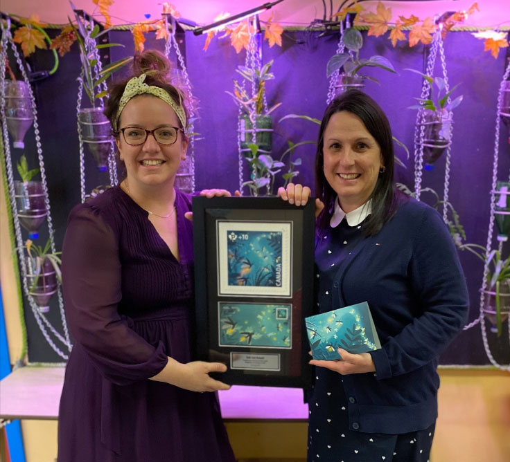 Teacher Sonia Demers and Principal Julie Guillemette of Saint-Romuald school hold a framed Community Foundation fundraising stamp and Official First Day Cover.